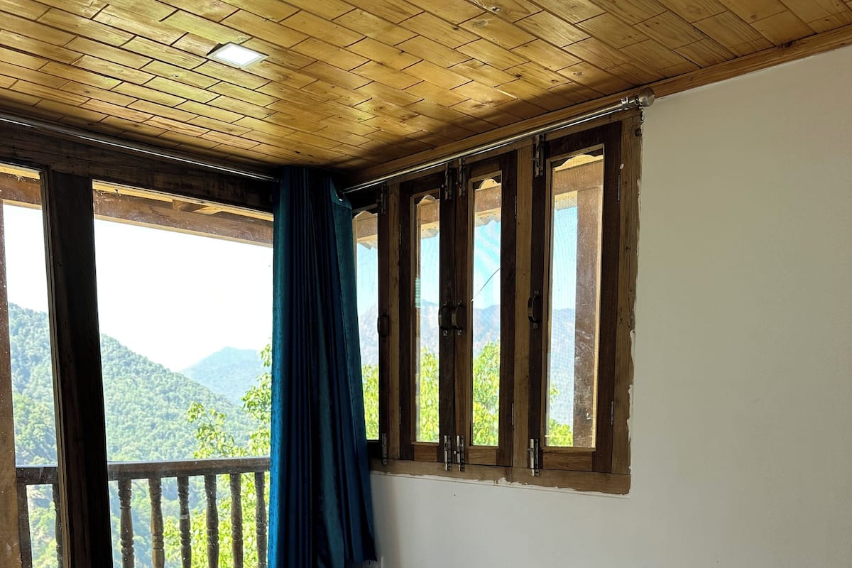 Un dormitorio con techo de madera, una cama grande con ropa blanca, dos toallas enrolladas, un balcón con cortina azul y vistas a colinas verdes.