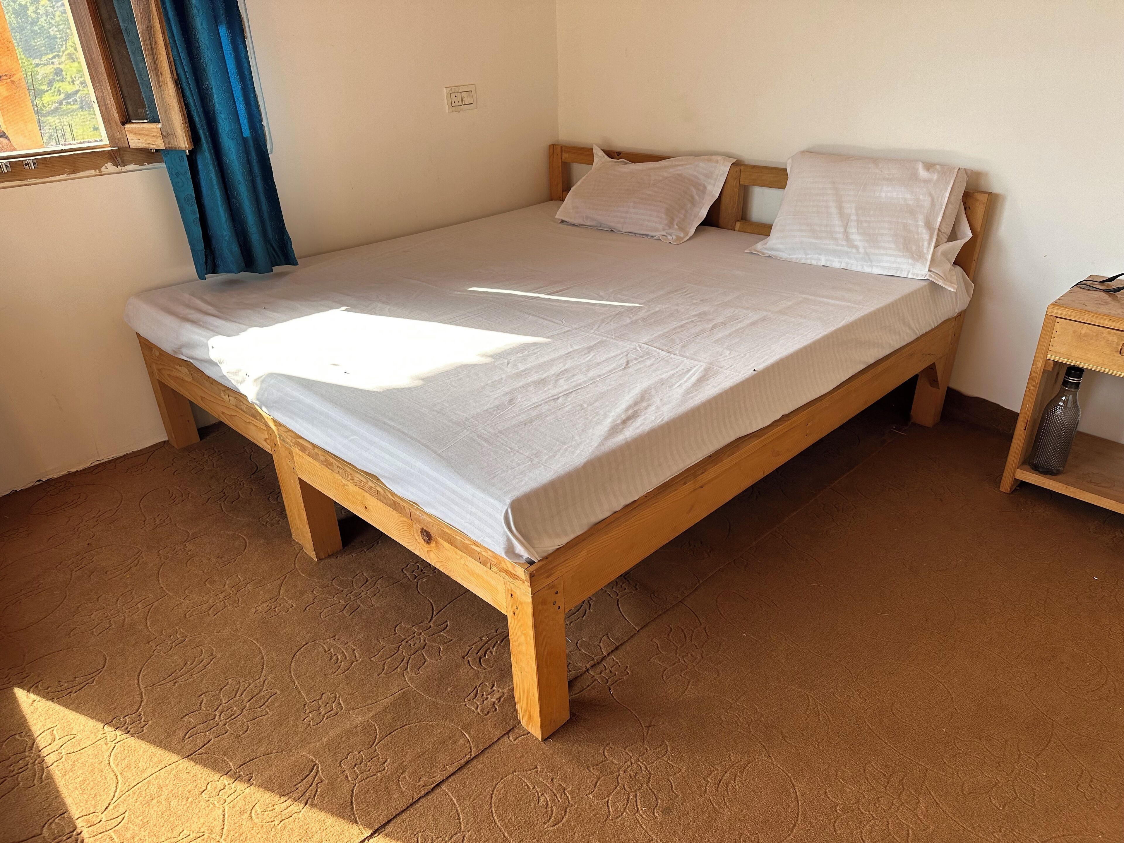 A single bed with a wooden frame and a white mattress, a wooden nightstand with a water bottle, and a window with blue curtains.
