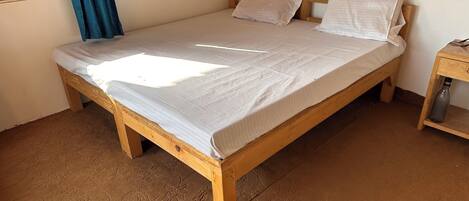 A single bed with a wooden frame and a white mattress, a wooden nightstand with a water bottle, and a window with blue curtains.