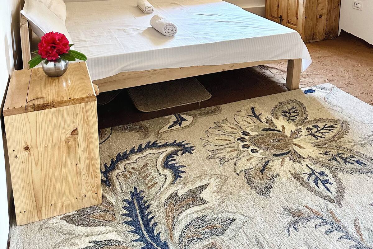 Un dormitorio con cama de madera, una mesita de noche con un florero y una ventana con contraventanas de madera, además de una alfombra estampada.
