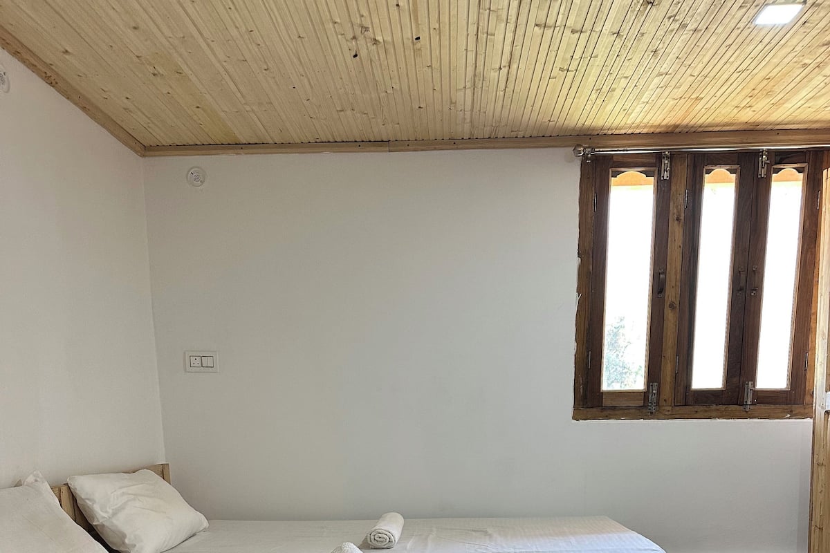 Una cama individual con ropa de cama blanca, un cabecero de madera y una toalla enrollada sobre la cama. Se ve una ventana con marcos de madera y vistas al exterior.