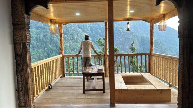Ein hölzerner Balkon mit einer Person, die auf eine gebirgige Landschaft blickt.