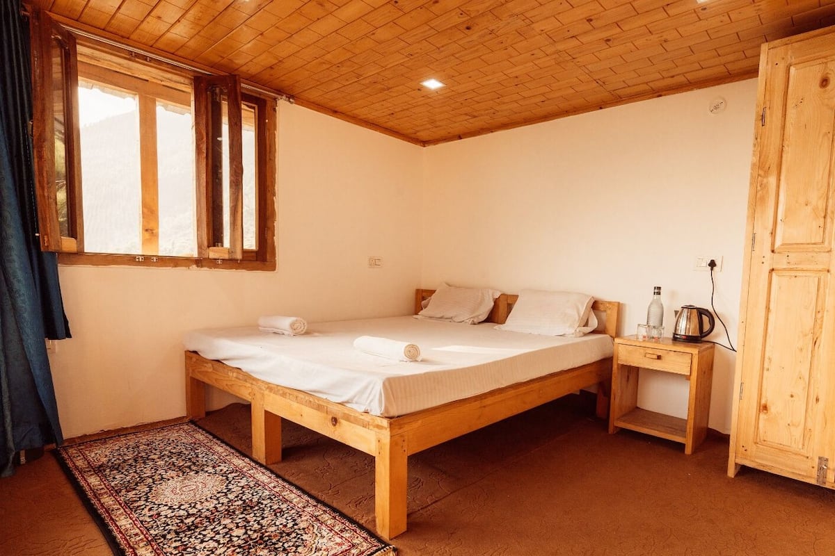 Una cama de madera con sábanas blancas, una mesita de noche con hervidor y un armario en una habitación con vigas de madera y una ventana.