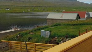 A lakeside view with a wooden deck, a small building with a red roof, and distant mountains under a cloudy sky.
