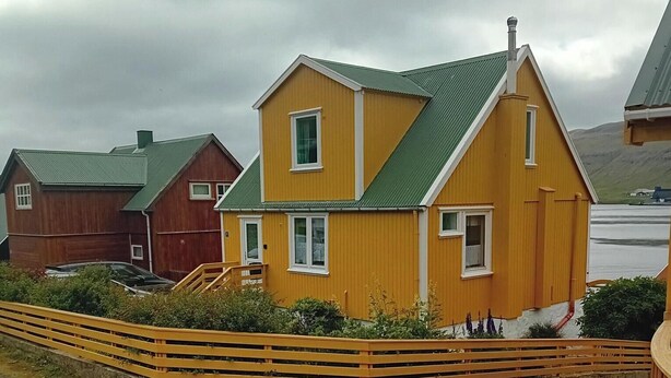 A yellow house with a green roof, surrounded by a wooden fence and greenery.