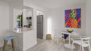A modern kitchen with white cabinetry, a stainless steel refrigerator, and a dining area with white chairs and a table. - Sandy Cove Hideaway: Private, End-Unit, Walk to Village, Private Beach, Fishing Pier & Dog Friendly! (Siesta Key)