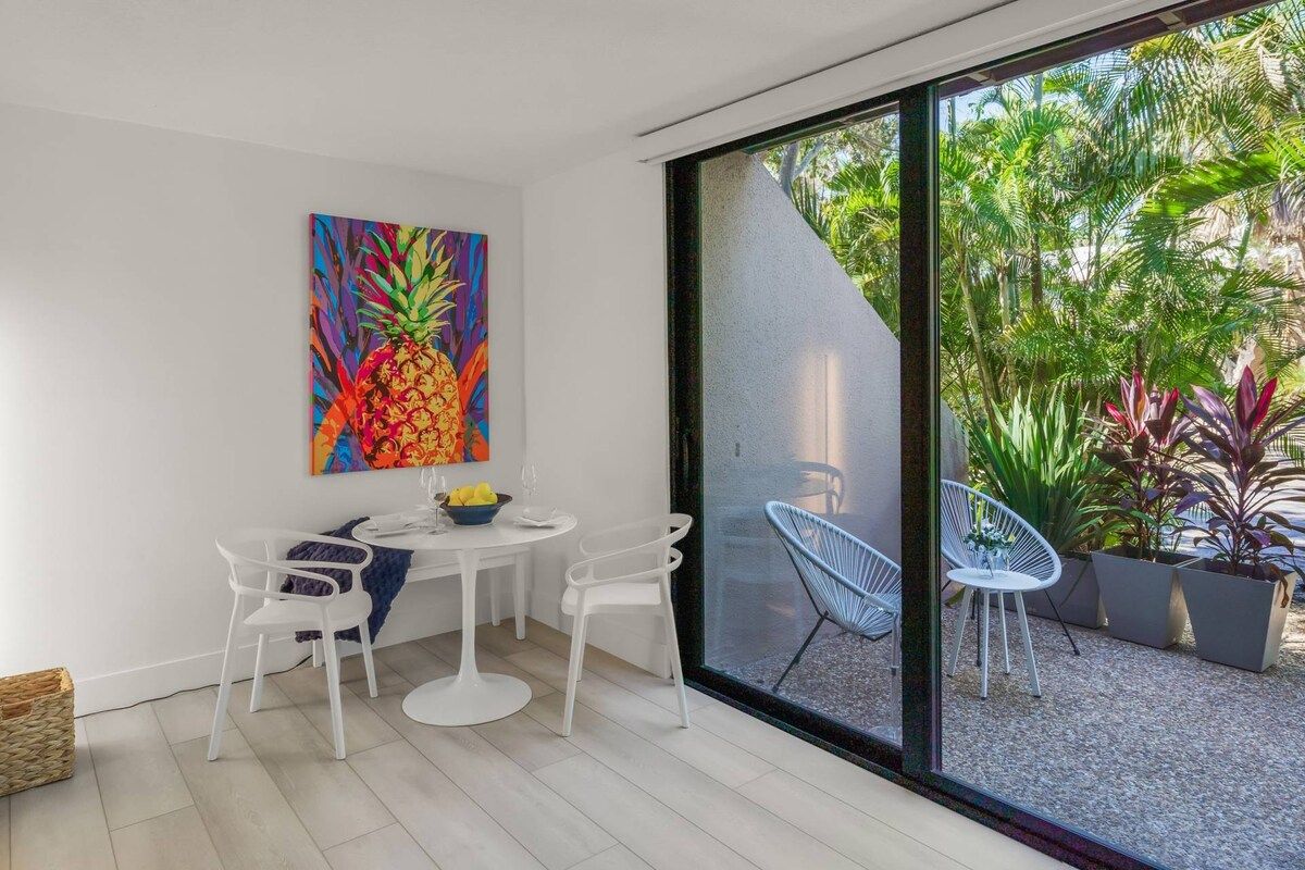 A modern dining area with a white table and chairs, a colorful pineapple painting on the wall, and a view of a garden through large glass doors.
