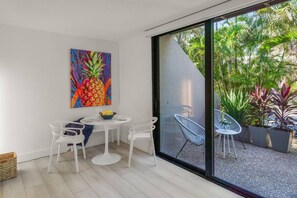 Ein moderner Essbereich mit einem weißen Tisch und Stühlen, einem farbenfrohen Ananas-Gemälde an der Wand und Blick auf einen Garten durch große Glasüren.
