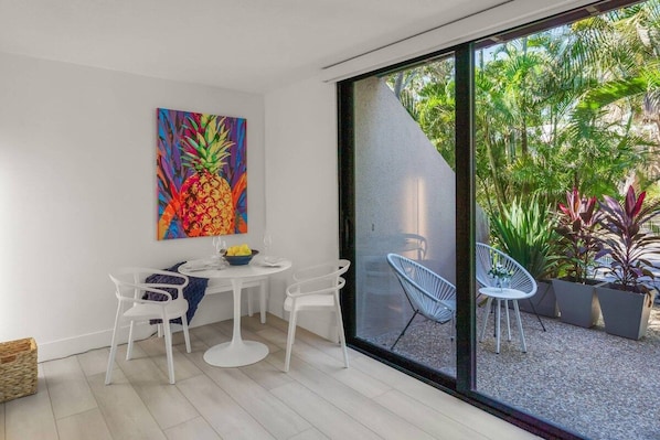 A modern dining area with a white table and chairs, a colorful pineapple painting on the wall, and a view of a garden through large glass doors.