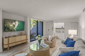 A modern living room with a flat-screen TV, a sofa, a coffee table, and a view of a balcony with outdoor furniture. - Secluded End Unit | Private Beach | Dog Friendly (Siesta Key)