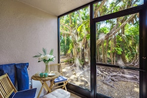 Une pièce dotée d’une grande fenêtre donnant sur une forêt tropicale. On y trouve une table en bois avec une chaise, une petite plante en pot et un coussin.