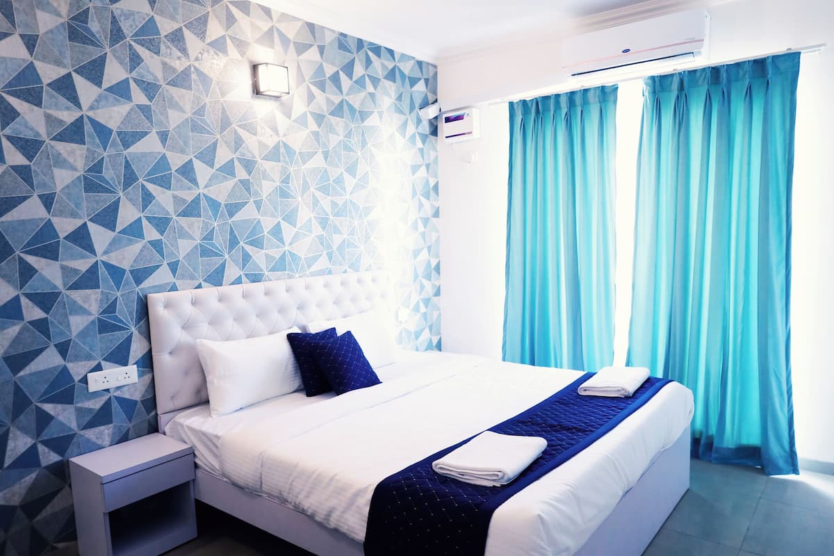 Une chambre moderne avec un mur orné d’un motif géométrique, un lit blanc agrémenté de touches de bleu, un ventilateur de plafond et une fenêtre avec des rideaux bleus.