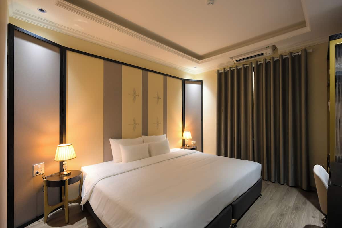 A hotel room with a large bed, two bedside tables with lamps, a wall-mounted air conditioner, and a curtain on the window.