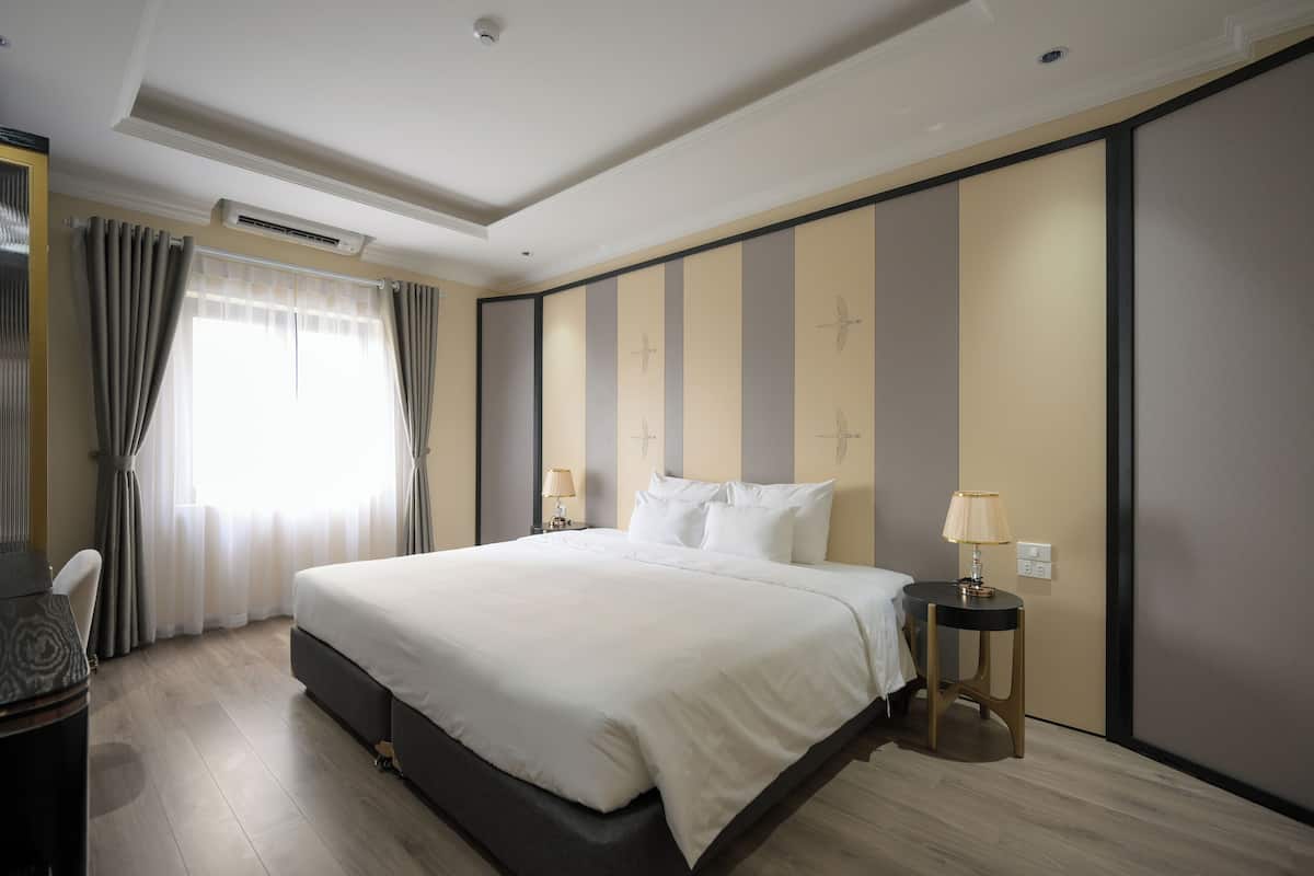 A modern hotel room with a large bed, a desk with a chair, a small table with a lamp, and a window with curtains.
