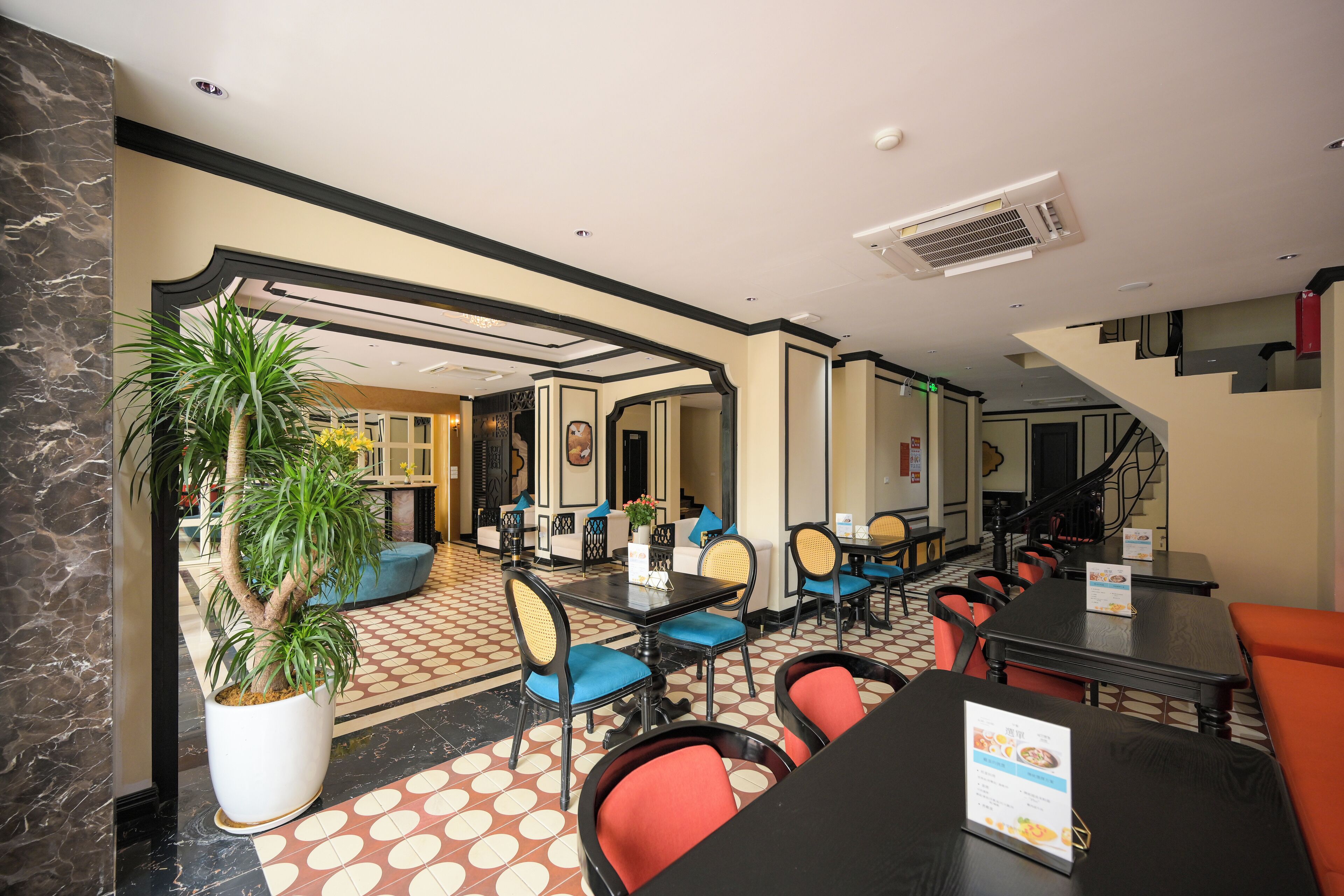 Ein Hotelrestaurant mit mehreren Tischen und Stühlen, einer großen Pflanze im Topf und einer Treppe im Hintergrund.