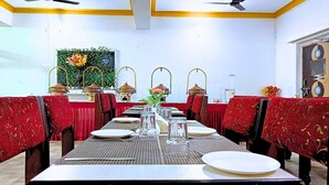 A dining area with red upholstered chairs, a long table set with plates, glasses, and a vase of flowers. - Hotel Vijay Deluxe (Sultanpur)