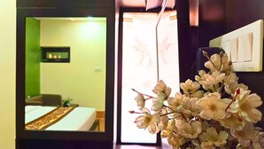 A room with a bed, a bedside table, and a mirror reflecting the room. There is a window with a view of the outside. - Hotel Vijay Deluxe (Sultanpur)