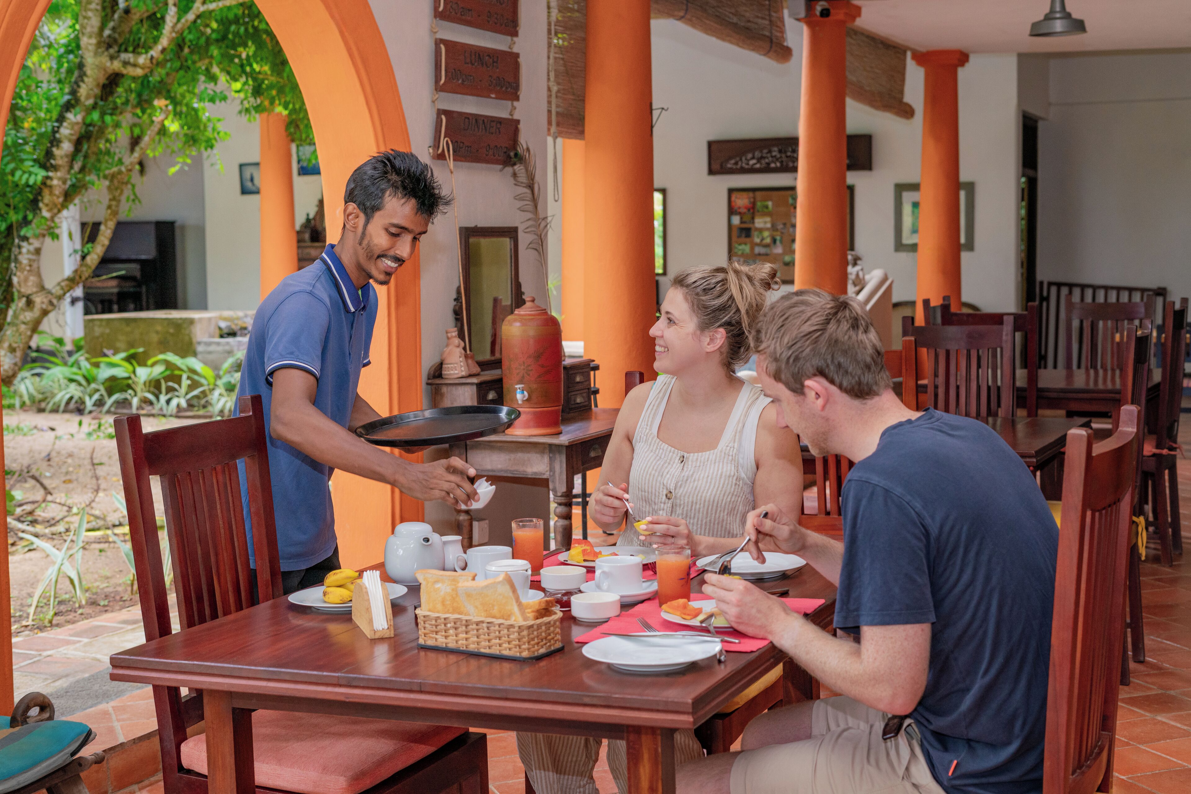 A dining area with wooden tables and chairs, a waiter serving food, and a tropical garden view.