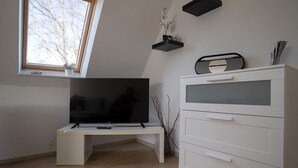 Moderni olohuone, jossa on valkoinen, litteänäyttöinen televisio valkoisella jalustalla, valkoinen lipasto laatikoilla, pieni hylly koriste-esineitä varten ja ikkuna, josta avautuu näkymä puita kohti.