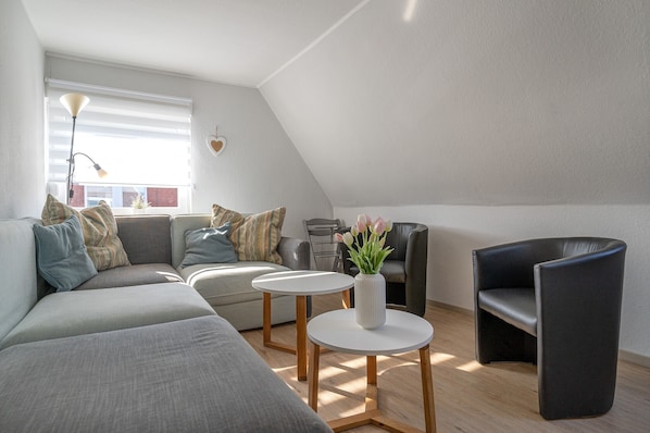 Un appartement moderne de style « loft » avec un plafond en pente, un canapé gris, un fauteuil en cuir noir et une petite table blanche agrémentée d’un vase de fleurs.