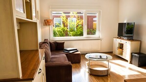 Ein modernes Wohnzimmer mit einer Couch, einem Hocker, einem Couchtisch, einem Fernseher und einem Fenster mit Blick auf Grünflächen.