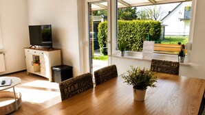 Ein Zimmer mit einem Holzesstisch, vier stuhligen Stühlen, einem Fernseher auf einer Kommode und Blick auf einen grünen Heckenstreifen.