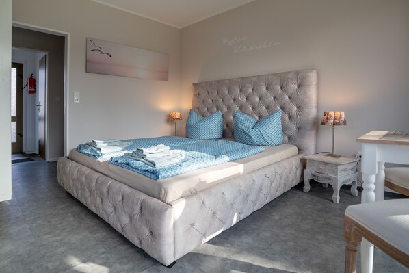 A bedroom with a tufted headboard, a bed with blue and white bedding, a bedside table with a lamp, and a small white nightstand. - Apartment 'Seestern' with Shared Garden and Wi-Fi (Norden)