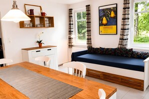 A modern room with a wooden dining table, white chairs, a sofa with a dark blue cushion, a small potted plant, a shelf with glassware, and a poster on the wall.