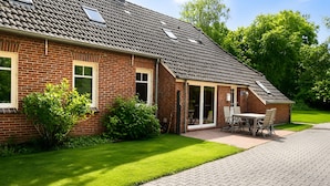 A brick house with a sloped roof, a covered patio with a table and chairs, and a well-maintained lawn. - Holiday Home 'Birken' with Shared Garden and Wi-Fi (Hage)