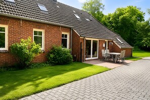 Ein Backsteinhaus mit geneigtem Dach, einer überdachten Terrasse mit Tisch und Stühlen sowie einem gepflegten Rasen.