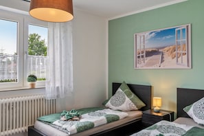 A bedroom with a view of the beach, a window with curtains, a radiator, and a bedside table with a lamp.