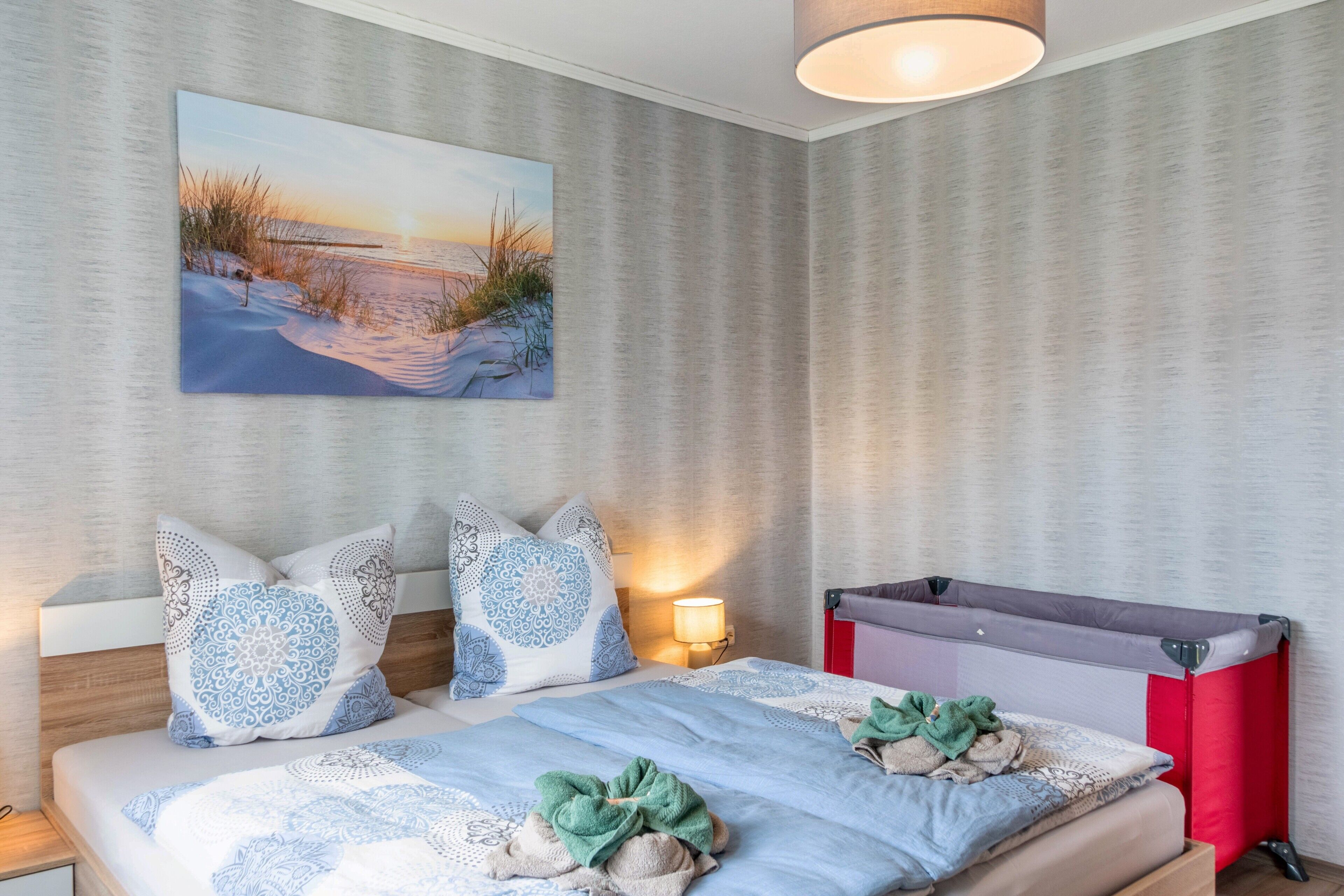A bedroom with a bed, pillows, a blanket, a painting, a lamp, and a crib.