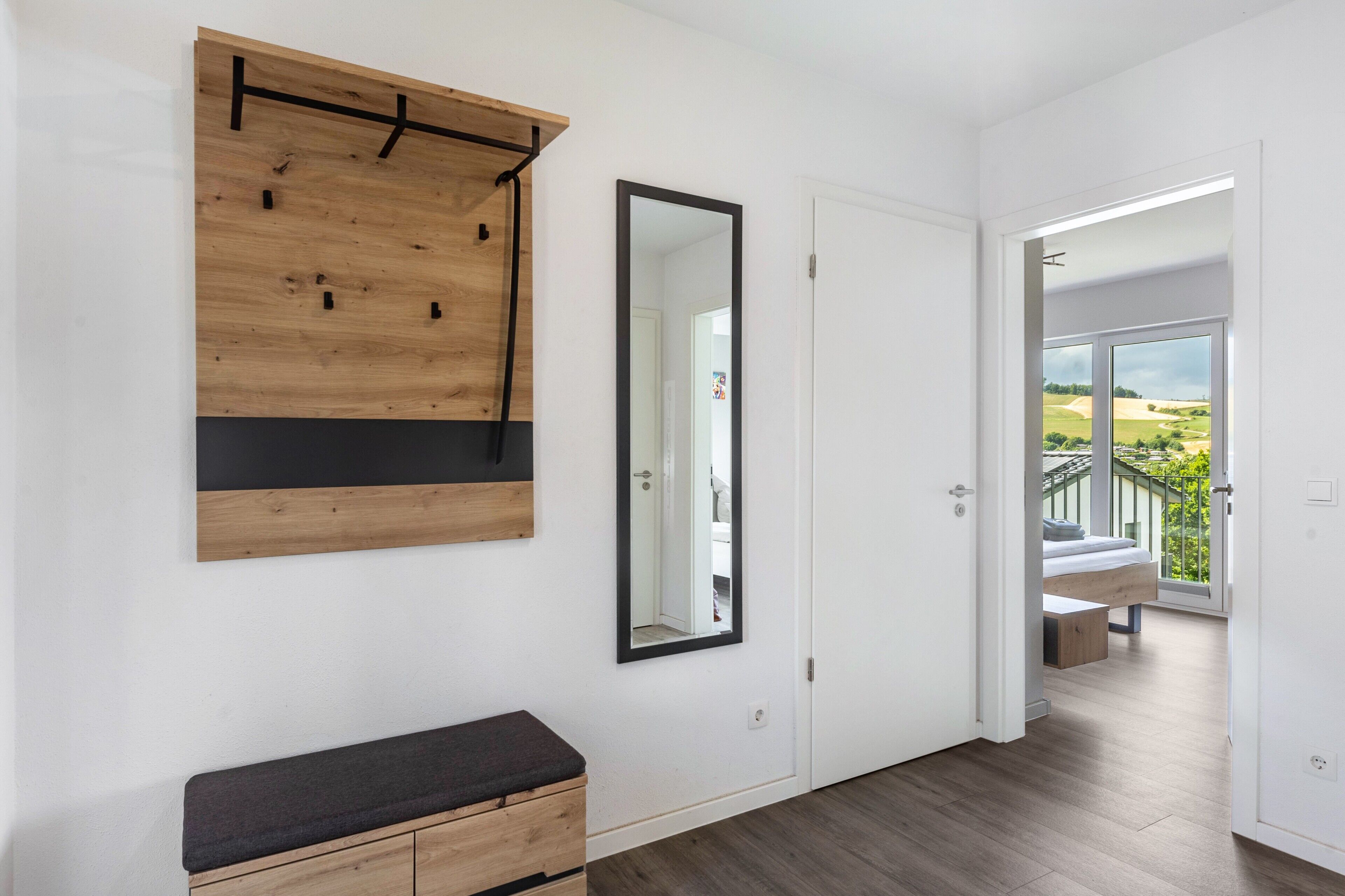 A modern room with a wooden wall-mounted shelf, a large mirror, a bench, and a door leading to a balcony with a view.