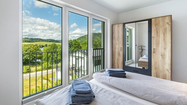 A bedroom with large windows, a bed, a wooden wardrobe, and a view of greenery. - Holiday Home 'Diemelblick' with Lake View, Private Terrace and Wi-Fi (Diemelsee)