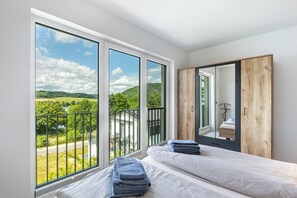 Et soveværelse med store vinduer, en seng, et træskab og udsigt til grønne omgivelser.