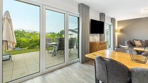 A modern hotel room with a large sliding glass door leading to an outdoor patio, a wooden dining table, and a flat-screen TV mounted on the wall. - Holiday Home 'Diemelblick' with Lake View, Private Terrace and Wi-Fi (Diemelsee)