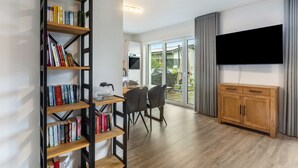 A modern living room with a bookshelf, a dining area, a TV mounted on the wall, and a sliding glass door leading to a garden. - Holiday Home 'Diemelblick' with Lake View, Private Terrace and Wi-Fi (Diemelsee)