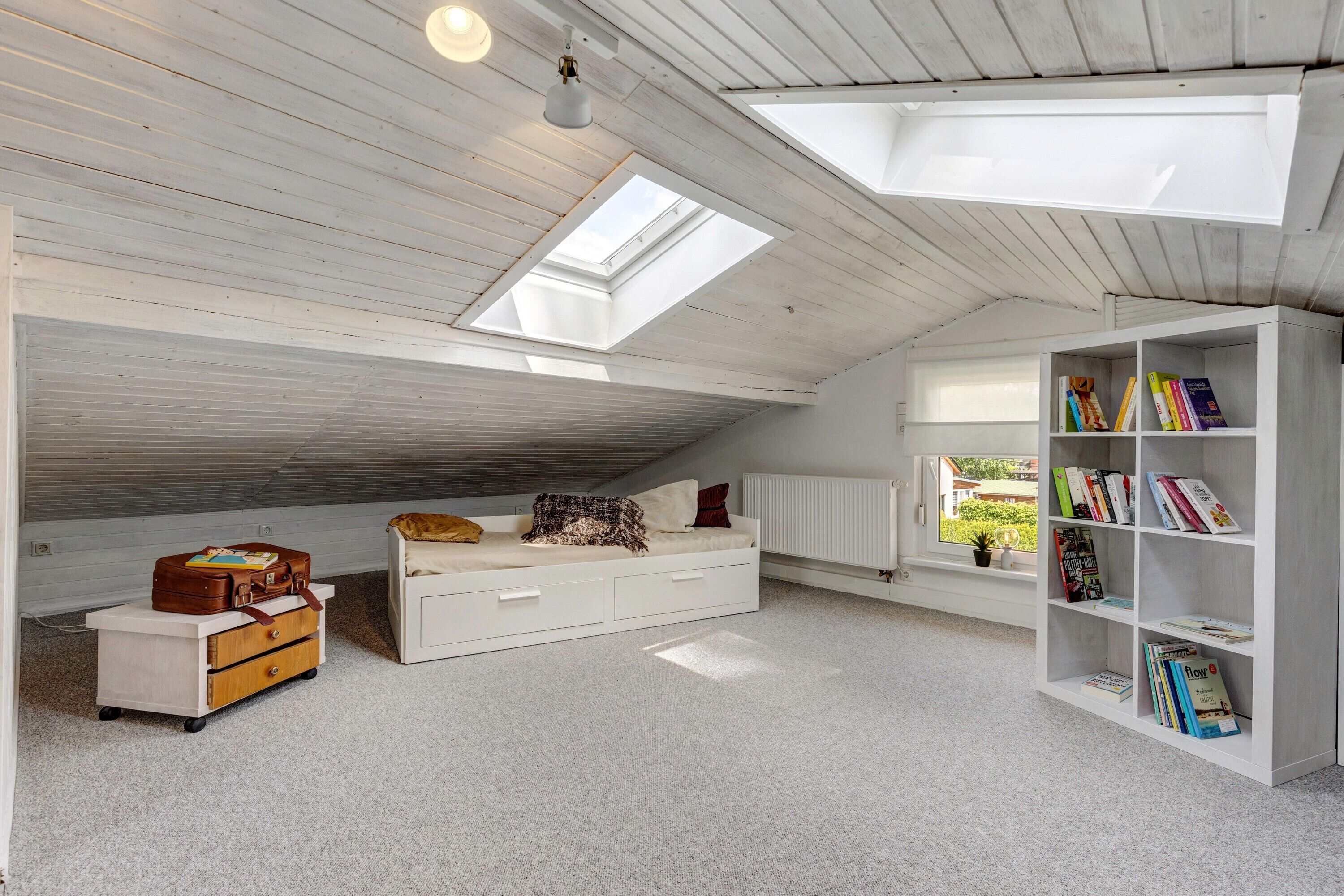 Una habitación con cama tipo altillo, cómoda blanca, estantería y tragaluces.