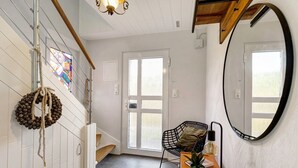 Une pièce avec un escalier en bois, un miroir rond, une chaise, une petite table avec une plante et un vitrail.