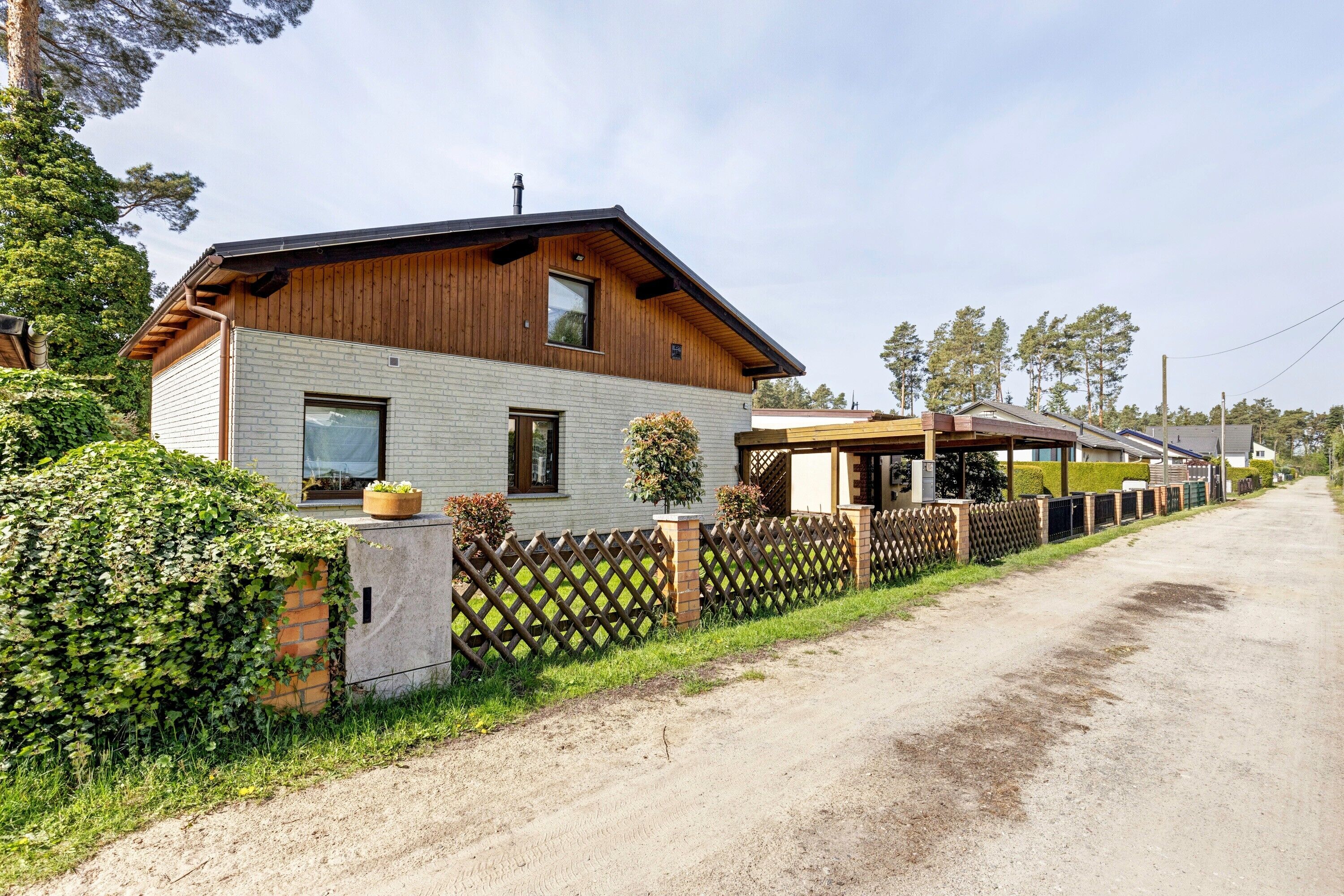 Une maison en bois à deux étages, avec des fondations en briques, une cour clôturée et un chemin de terre.
