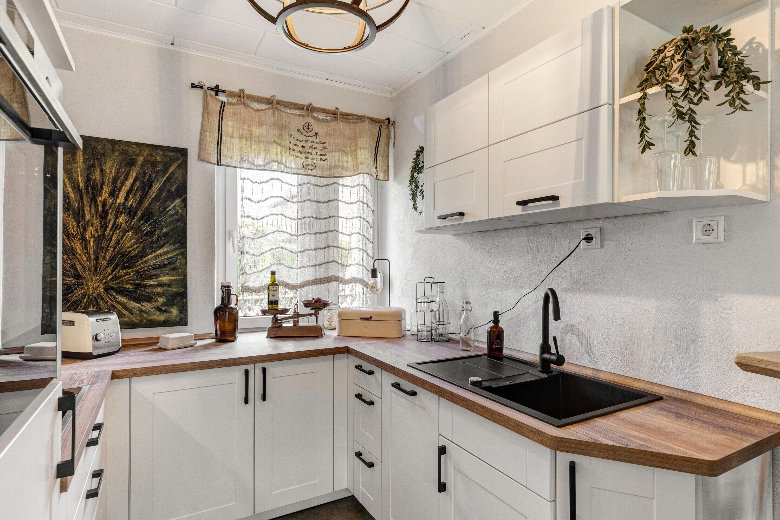 Een moderne keuken met witte kastjes, een houten aanrecht, een zwarte spoelbak en een raam met gordijn.
