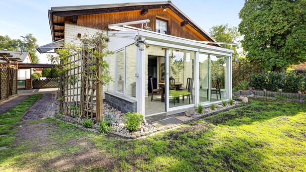 Une maison moderne dotée d’un patio entièrement vitré, d’une terrasse en bois et d’un chemin de pierre menant à l’entrée.