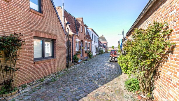 Een smalle straat met keien, geflankeerd door bakstenen gebouwen, met een rood voertuig aan de rechterkant en beplanting langs de randen.