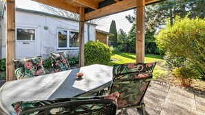 Een terras buiten met een tafel en stoelen, een houten pergola en uitzicht op een huis en tuin.