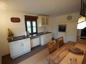 Une cuisine avec des meubles blancs, un plan de travail en bois, une table à manger avec des chaises, une horloge au mur et une fenêtre donnant sur des arbres.