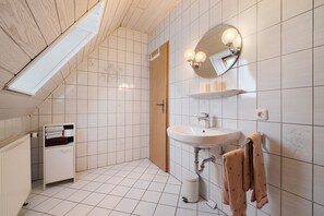 Moderni kylpyhuone, jossa on kattovalokuvio, valkoiset laatat, puuovi, valkoinen pesuallas ja pyyhekoukku.