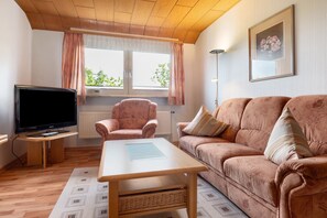 A living room with a television, a sofa, a chair, a table, and a window with curtains. - Apartment 'Monika' in Butjadingen (Butjadingen)