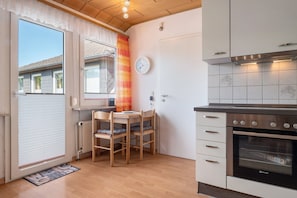 Ett kompakt kök med en inbyggd ugn, ett matbord med stolar, ett fönster med gardiner och en väggklocka.