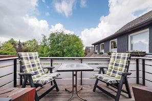 En balkong med två stolar och ett bord, omgiven av ett vitt räcke, med utsikt över ett bostadsområde med träd och ett hus.