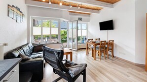 A modern living room with a wooden ceiling, a black leather sofa, a dining table with chairs, and a flat-screen TV mounted on the wall. - Holiday Home Little Lighthouse-Backbord Terrace, Garden, Wi-Fi, Bike Garage, EV Charging (Fehmarn)